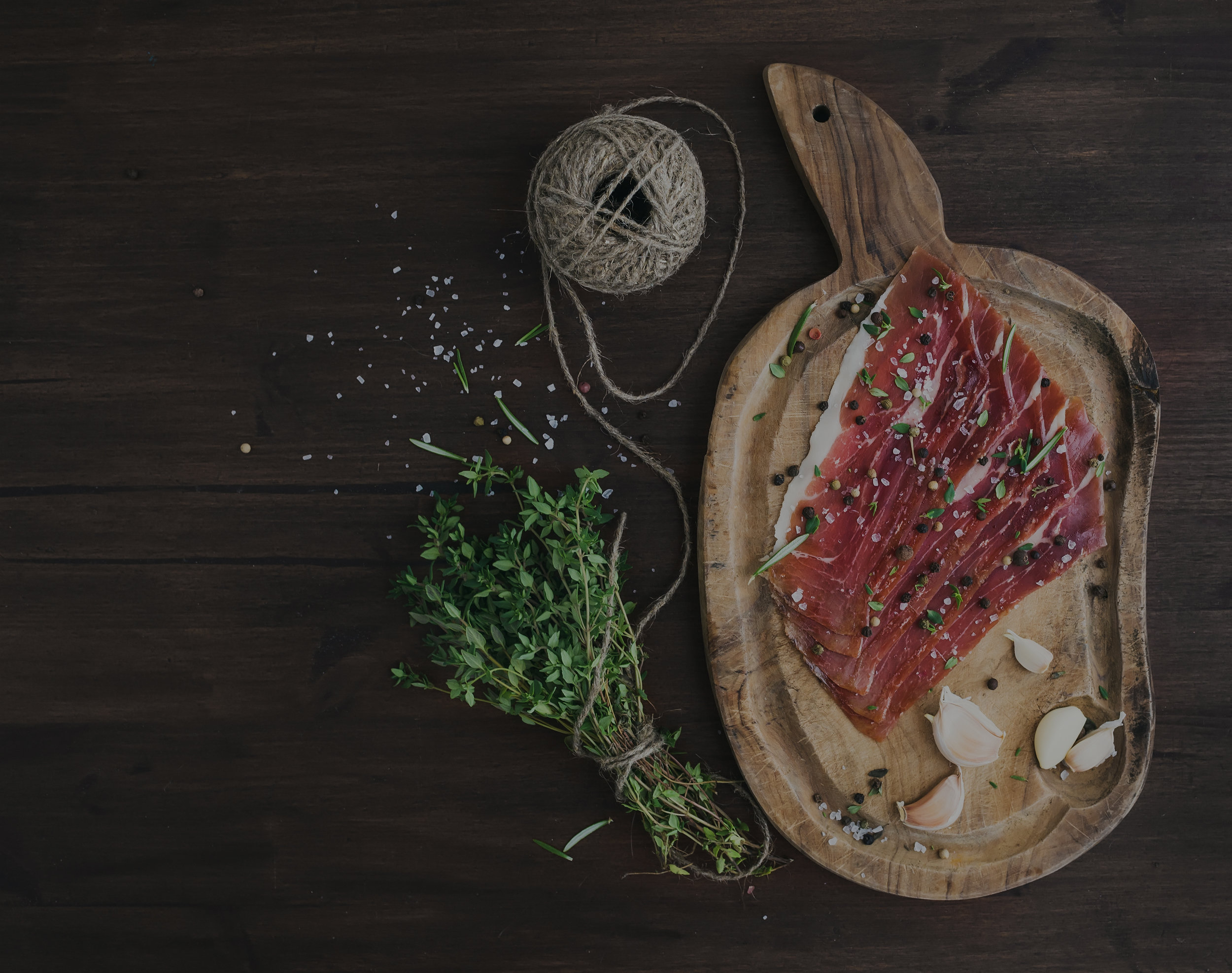 Cured pork meat or prosciutto on a rustic wooden board with garlic, spices and thyme over a dark wood background with a copy space. Top view